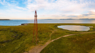 Et høyt, rødt metalltårn står på en gresslette i nærheten av et lite tjern og strandlinjen, med fjerne åser og en delvis overskyet himmel i bakgrunnen.