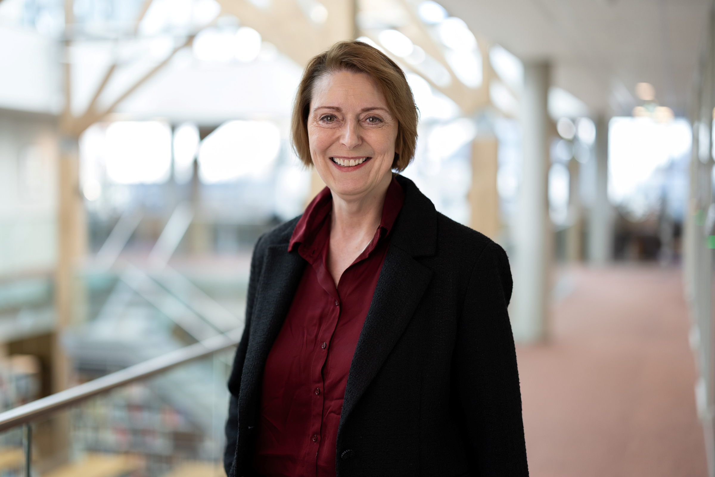 En kvinne med kort, lysebrunt hår, iført mørk blazer og rødbrun skjorte, står smilende i en lys innendørs gang med store vinduer og uskarp bakgrunn.
