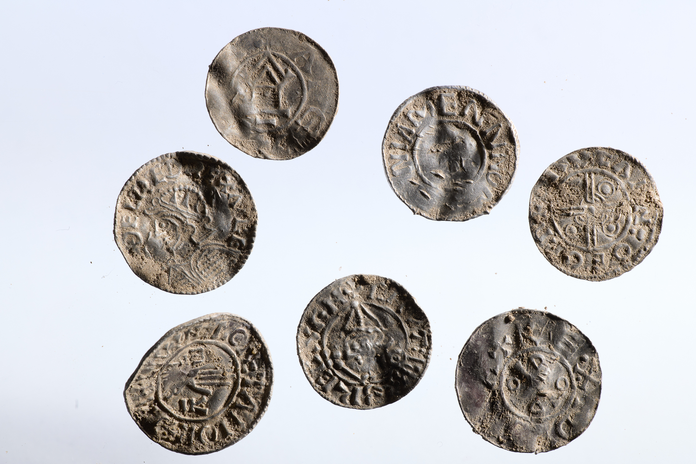 Sju gamle, forvitrede mynter med slitte graveringer og inskripsjoner er ordnet i et løst sirkelmønster på en vanlig hvit bakgrunn.