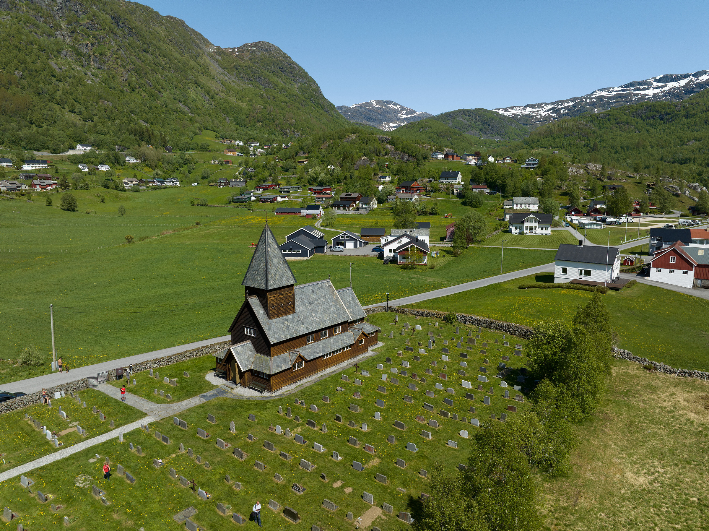 Flyfoto av en trekirke omgitt av en kirkegård med gravsteiner, i en grønn dal med spredte hus og fjell i bakgrunnen.