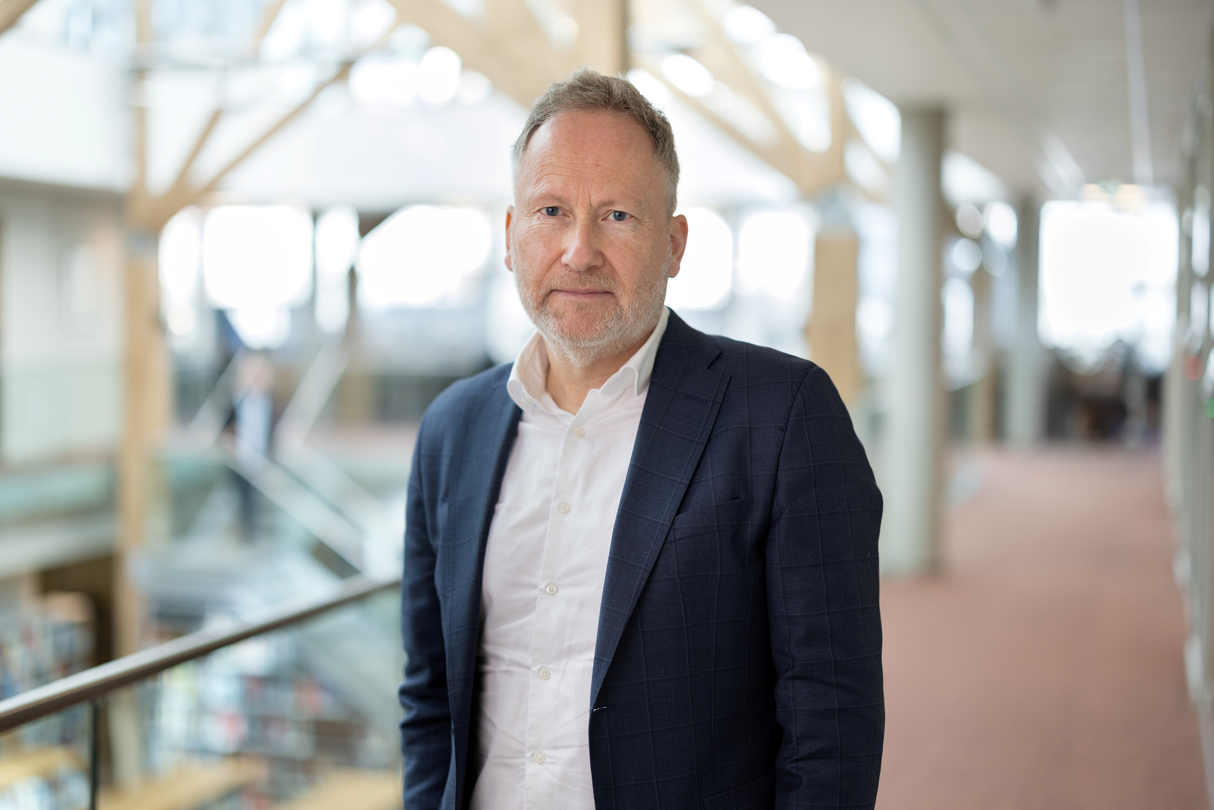 En mann i dress står i en lys korridor med glassrekkverk og en uskarp bakgrunn av vinduer og trebjelker.