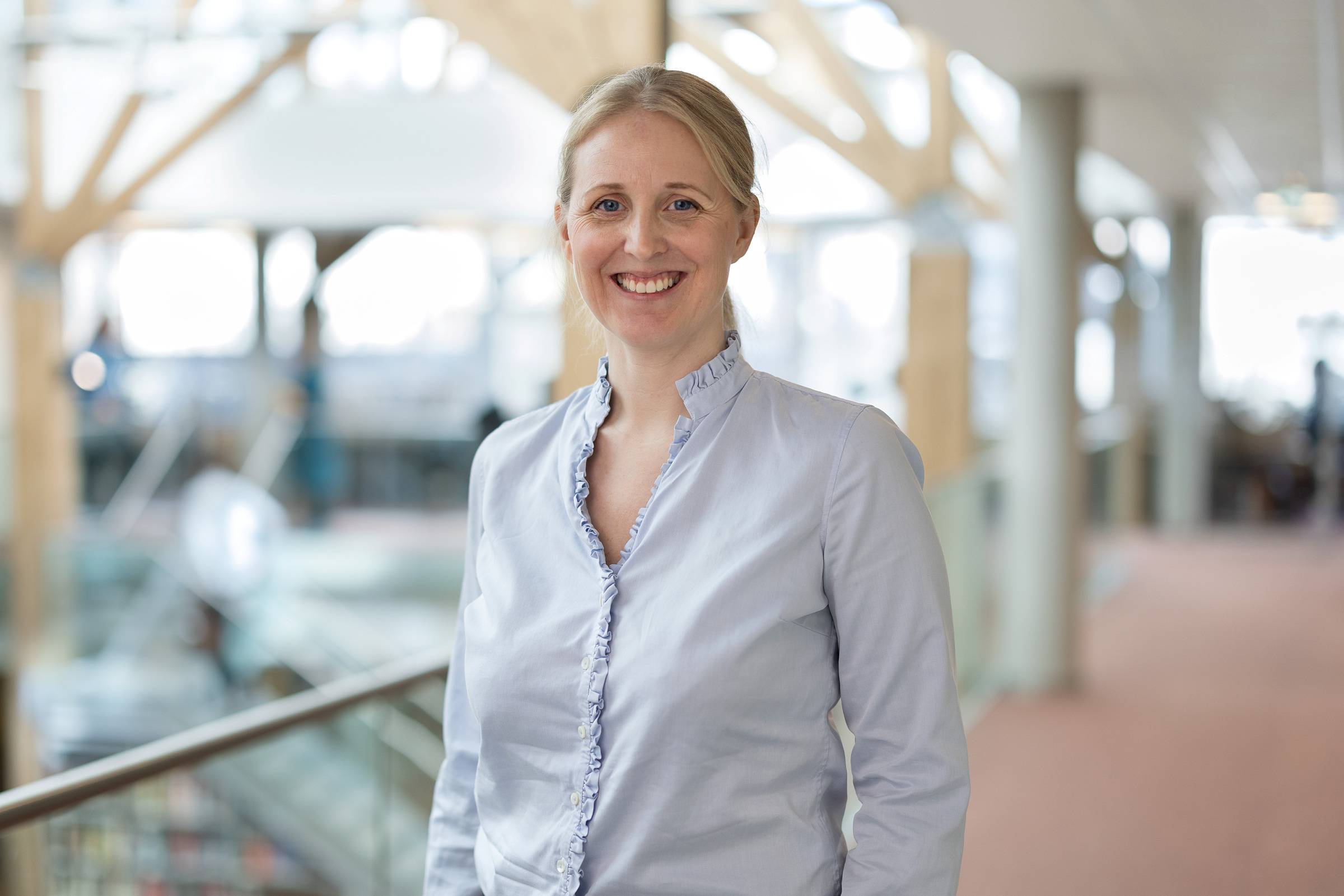 En kvinne i lyseblå bluse står innendørs og smiler, med en uskarp bakgrunn av et moderne kontor eller atrium.