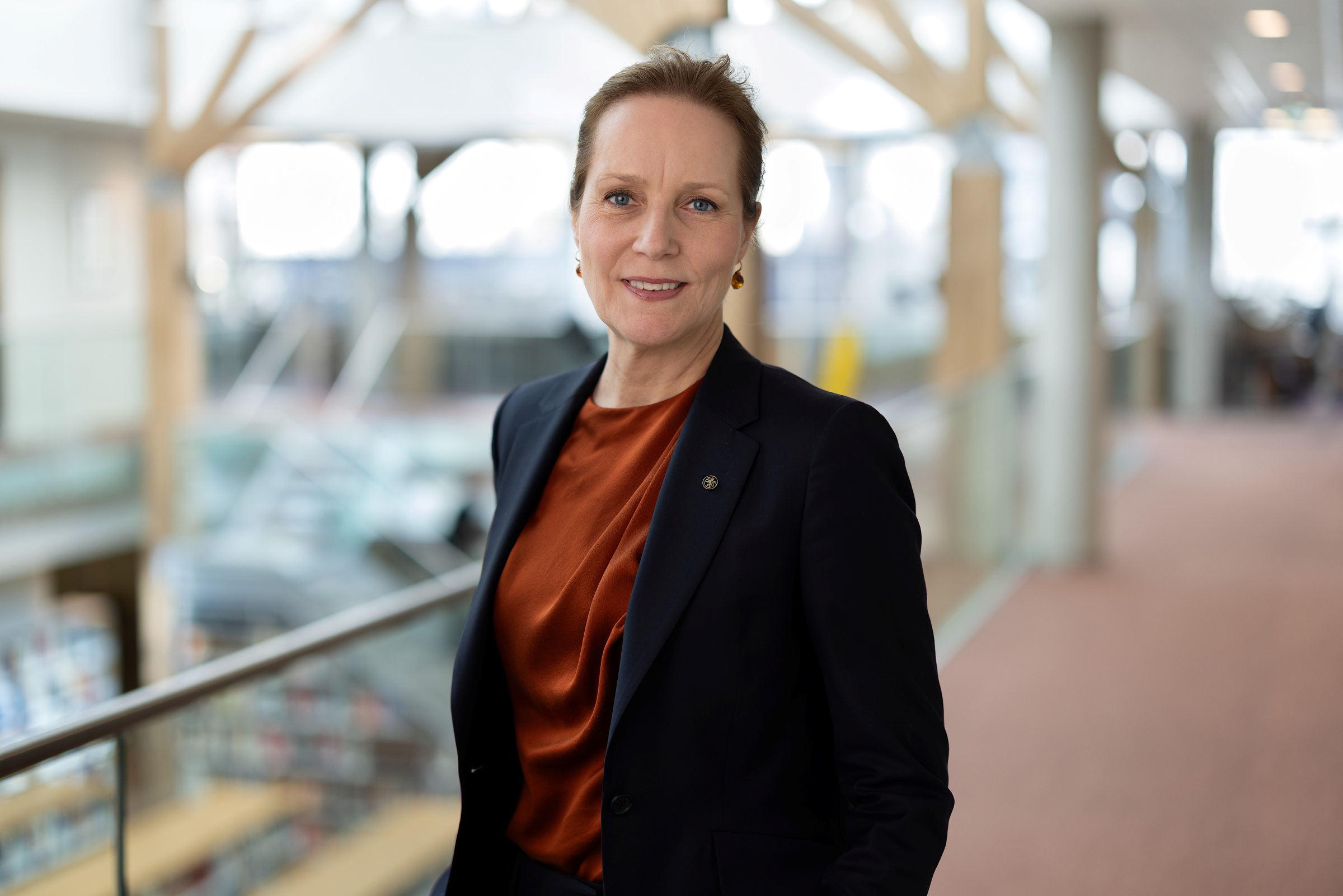 En kvinne i mørk blazer og rustfarget bluse står innendørs og smiler til kameraet i en moderne, lys bygning med glass- og tredetaljer.