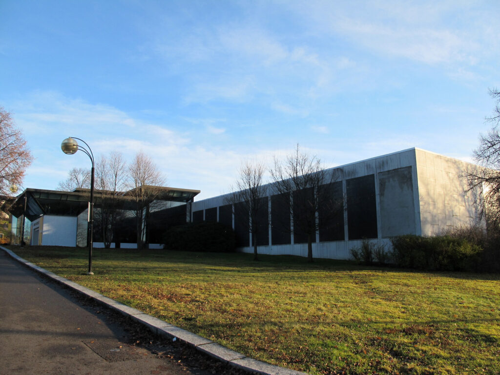 En moderne, rektangulær bygning med store vinduer og betongvegger ligger ved siden av en gressplen under en skyfri himmel.