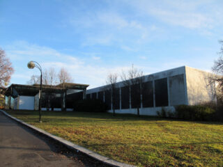 En moderne, rektangulær bygning med store vinduer og betongvegger ligger ved siden av en gressplen under en skyfri himmel.