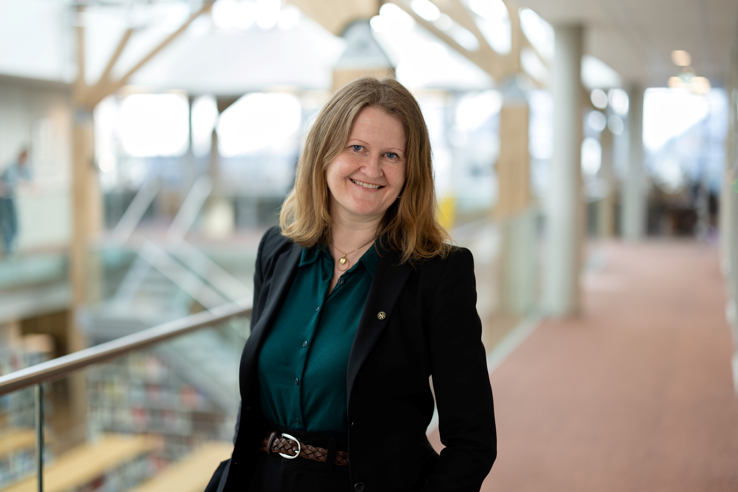 En kvinne med skulderlangt hår, iført mørk blazer og grønn bluse, står innendørs i en godt opplyst gang og smiler til kameraet.