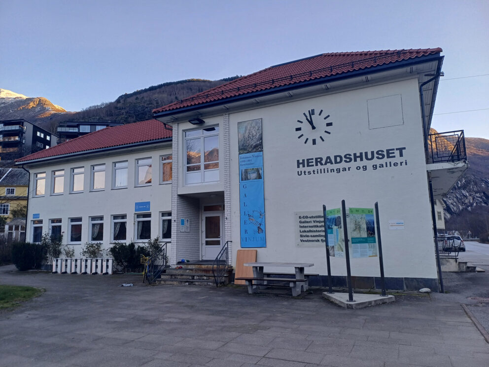 Heradshuset ligger i en hvit toetasjes bygning med rødt tegltak, med utstillings- og galleriskilt på norsk, utebenker og fjellene i bakgrunnen.