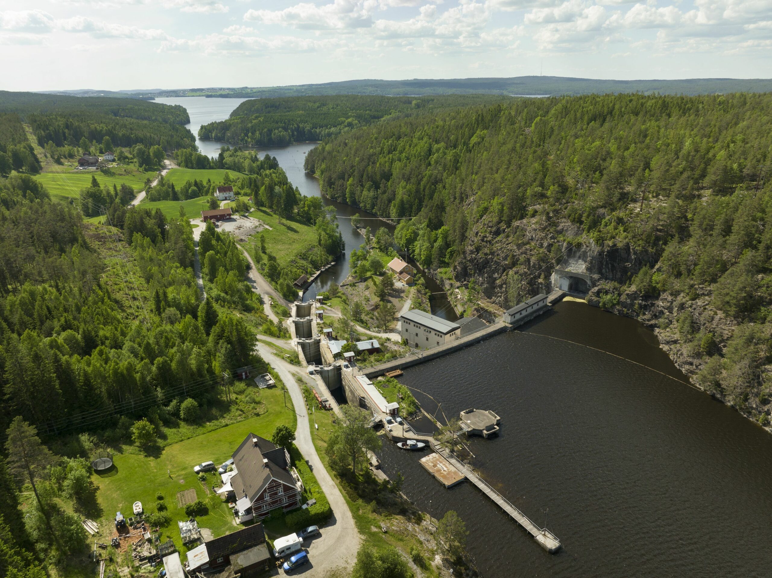 Flyfoto av et ruralt landskap med en vannkraftdam, elv, skogkledde åser, hus og klar himmel.