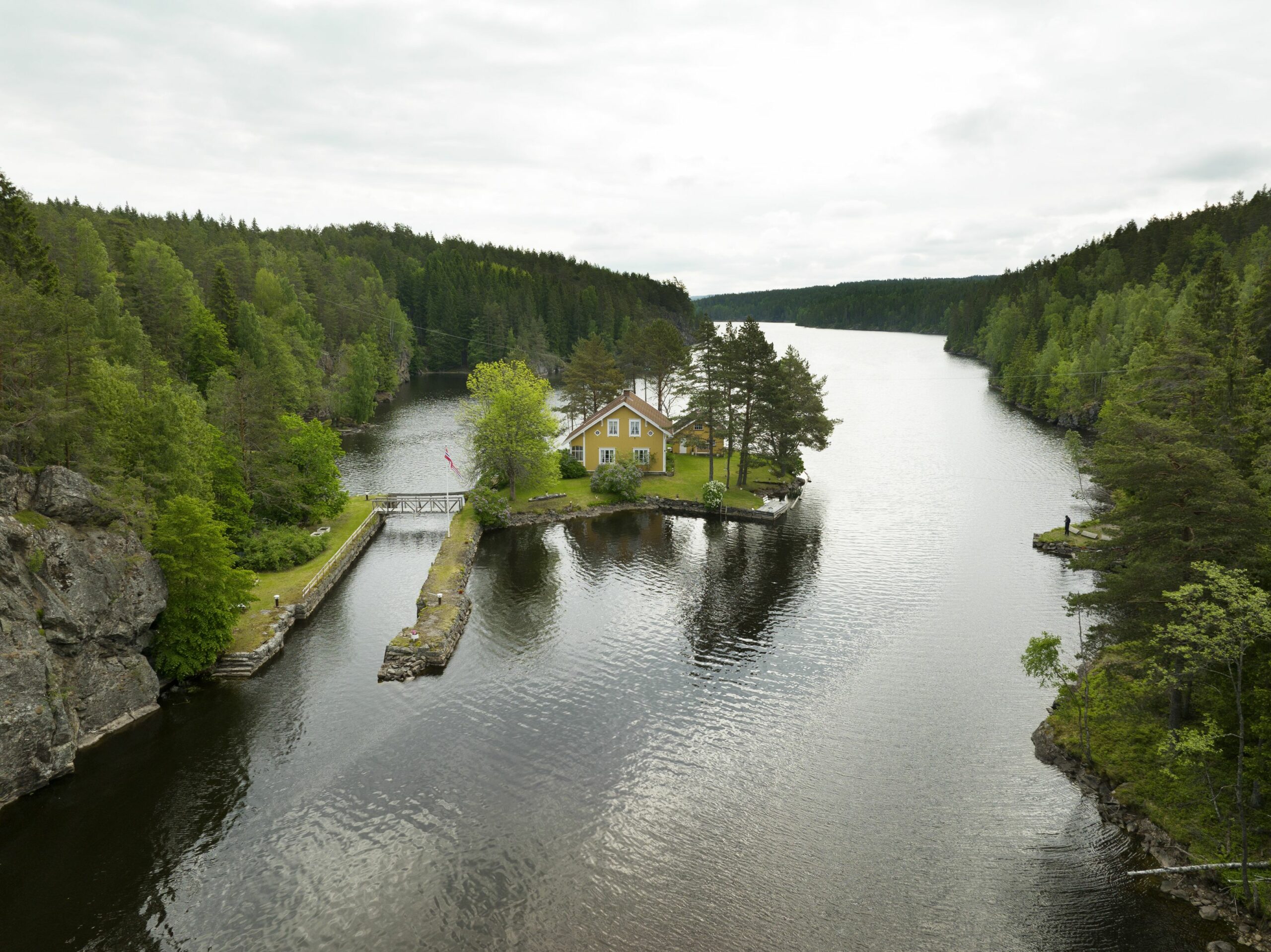 Et gult hus ligger på en liten halvøy omgitt av en stille innsjø, med tett, grønn skog på begge sider under en overskyet himmel.