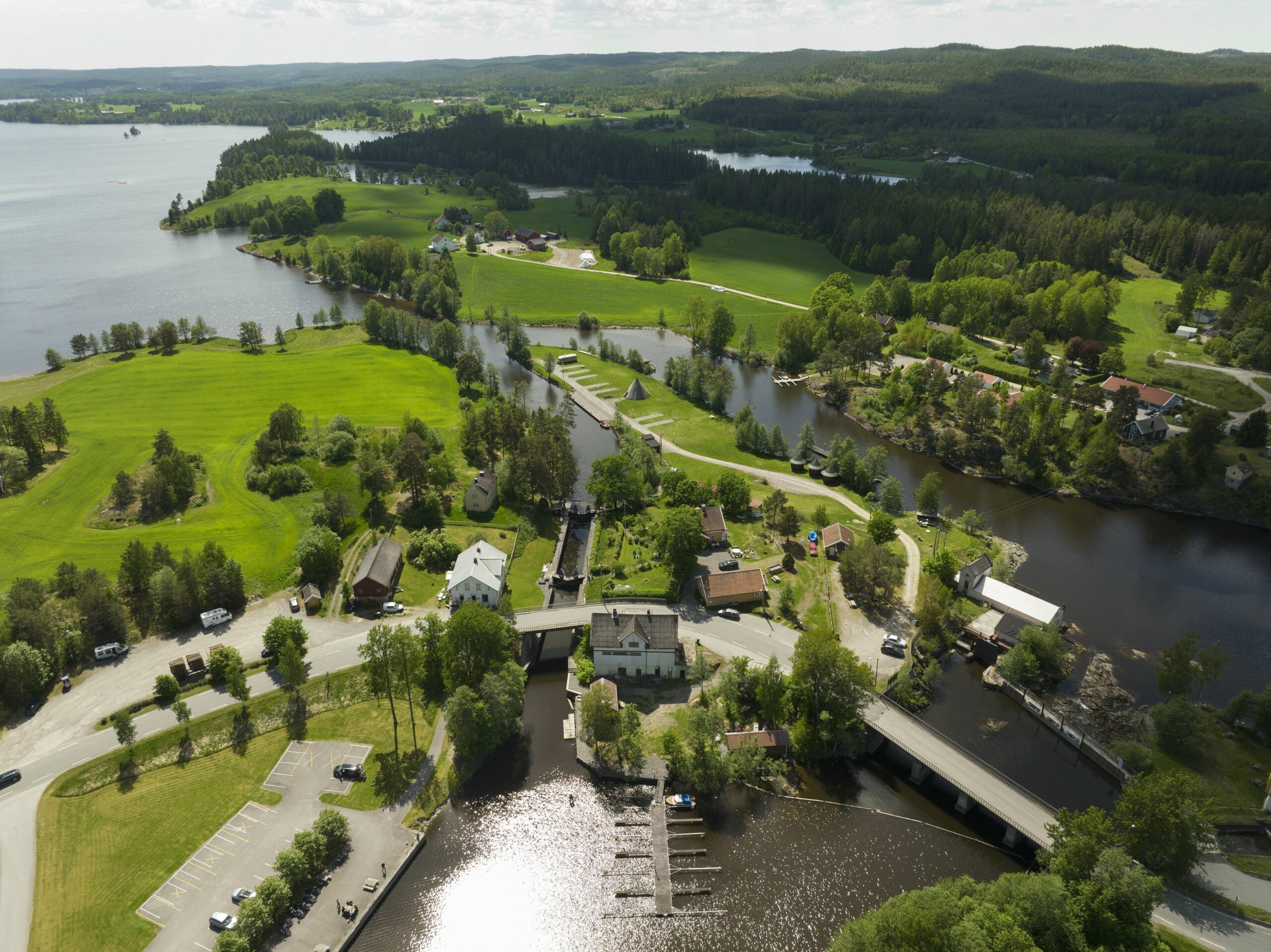 Flyfoto av et landlig landskap med en elv, grønne åkrer, spredte hus, en bro og båtbrygger nær en parkeringsplass, omgitt av trær og åser.