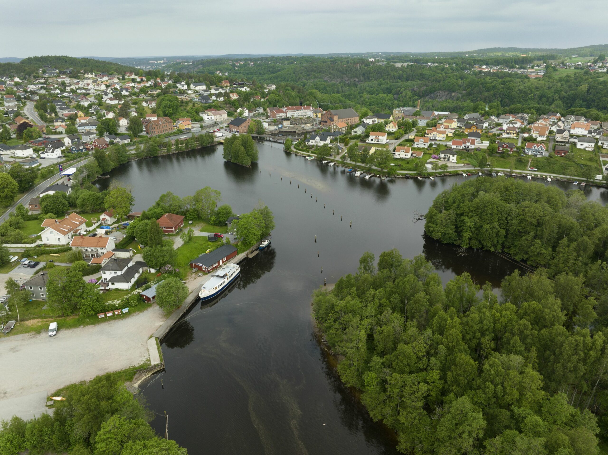 Flyfoto av en liten by med hus, trær og en elv med en båt som ligger til kai, omgitt av grøntområder under en overskyet himmel.
