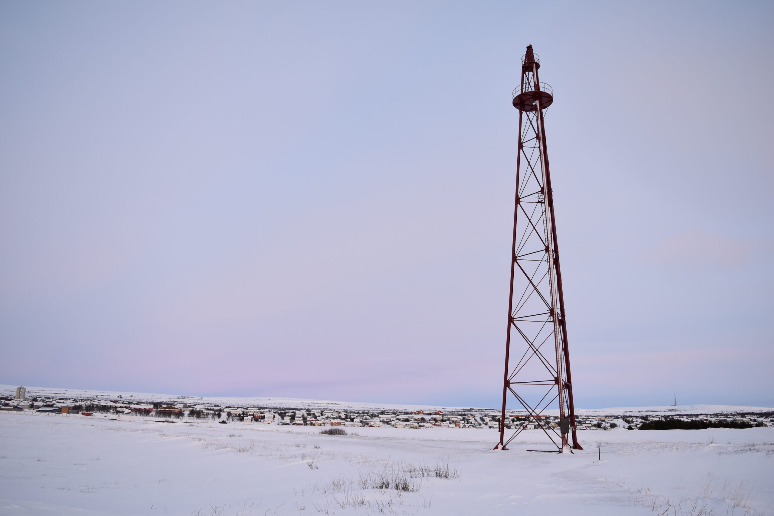 Et høyt metalltårn står i et snøkledd, flatt landskap med bygninger synlige i bakgrunnen under en blek himmel.