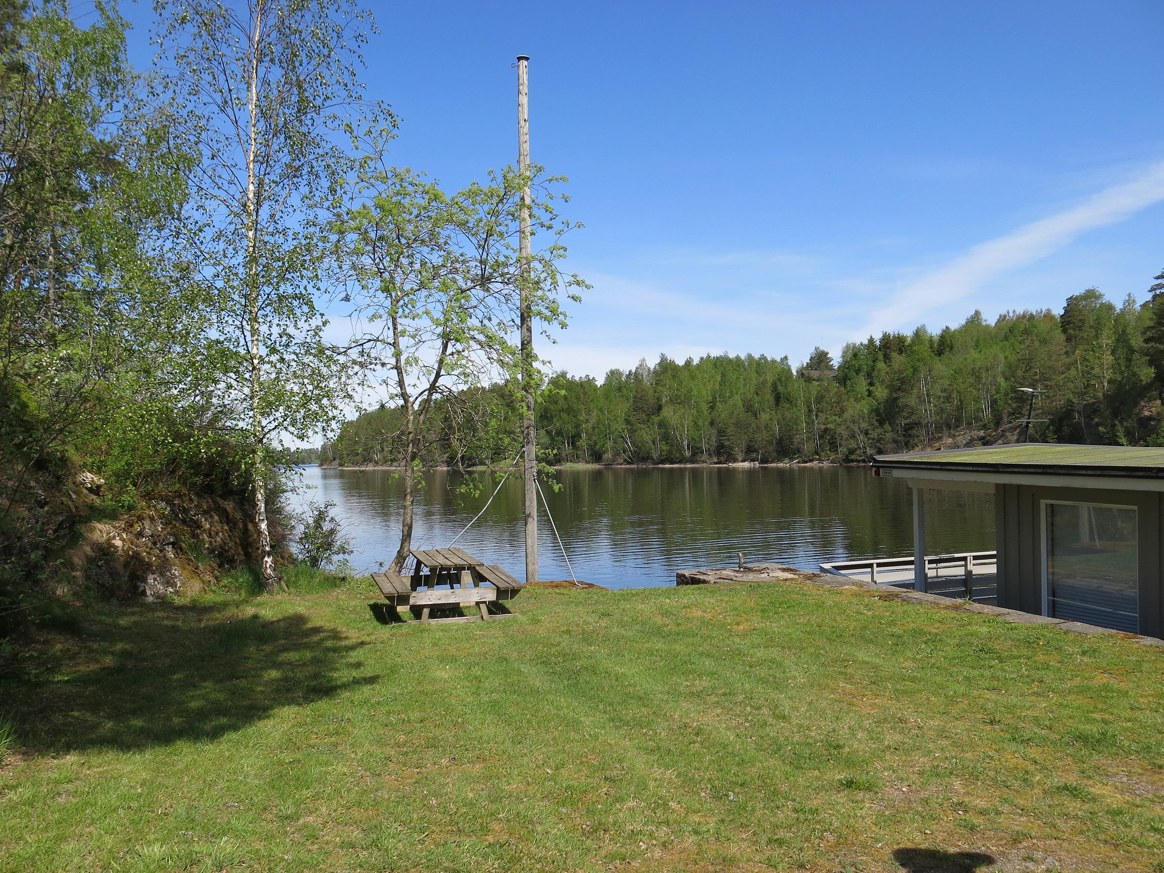 Fra et gressområde med piknikbord, trær og en liten brygge har du utsikt over en rolig innsjø omgitt av skog under en klarblå himmel.