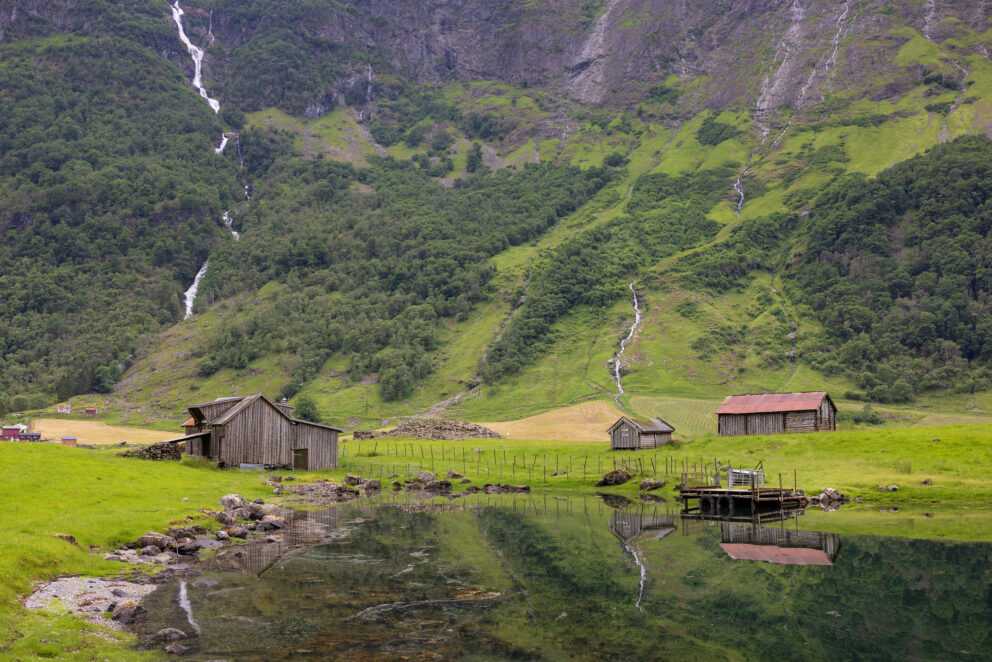 Tre trehytter står ved siden av et stille tjern som speiler dem, mot grønne åser med en liten foss og en overskyet himmel i bakgrunnen.