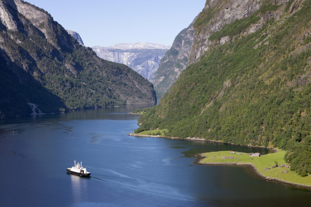 En hvit ferge seiler gjennom en smal, stille fjord omkranset av bratte, grønne fjell under en klar himmel.