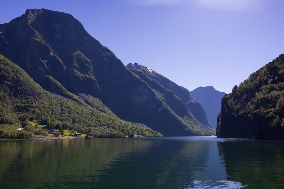 En rolig fjord omgitt av bratte, grønne fjell under en klarblå himmel, med synlige refleksjoner i vannoverflaten.