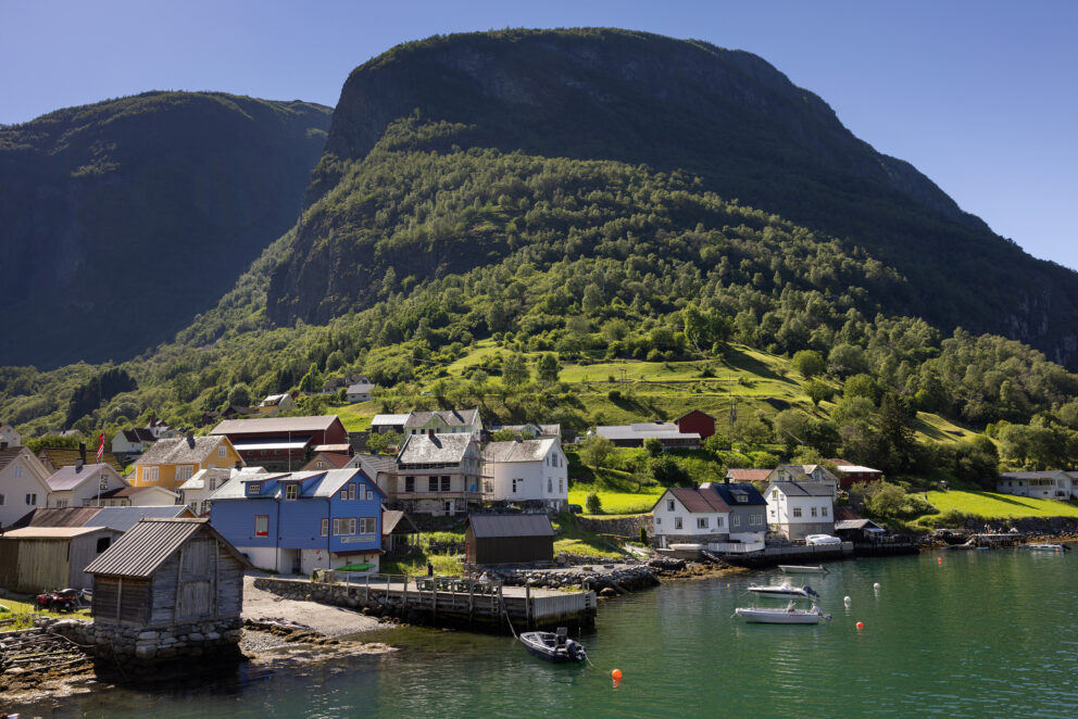 Liten kystlandsby med båter som ligger til kai ved vannet, tradisjonelle hus og et stort, grønt fjell i bakgrunnen under en klarblå himmel.