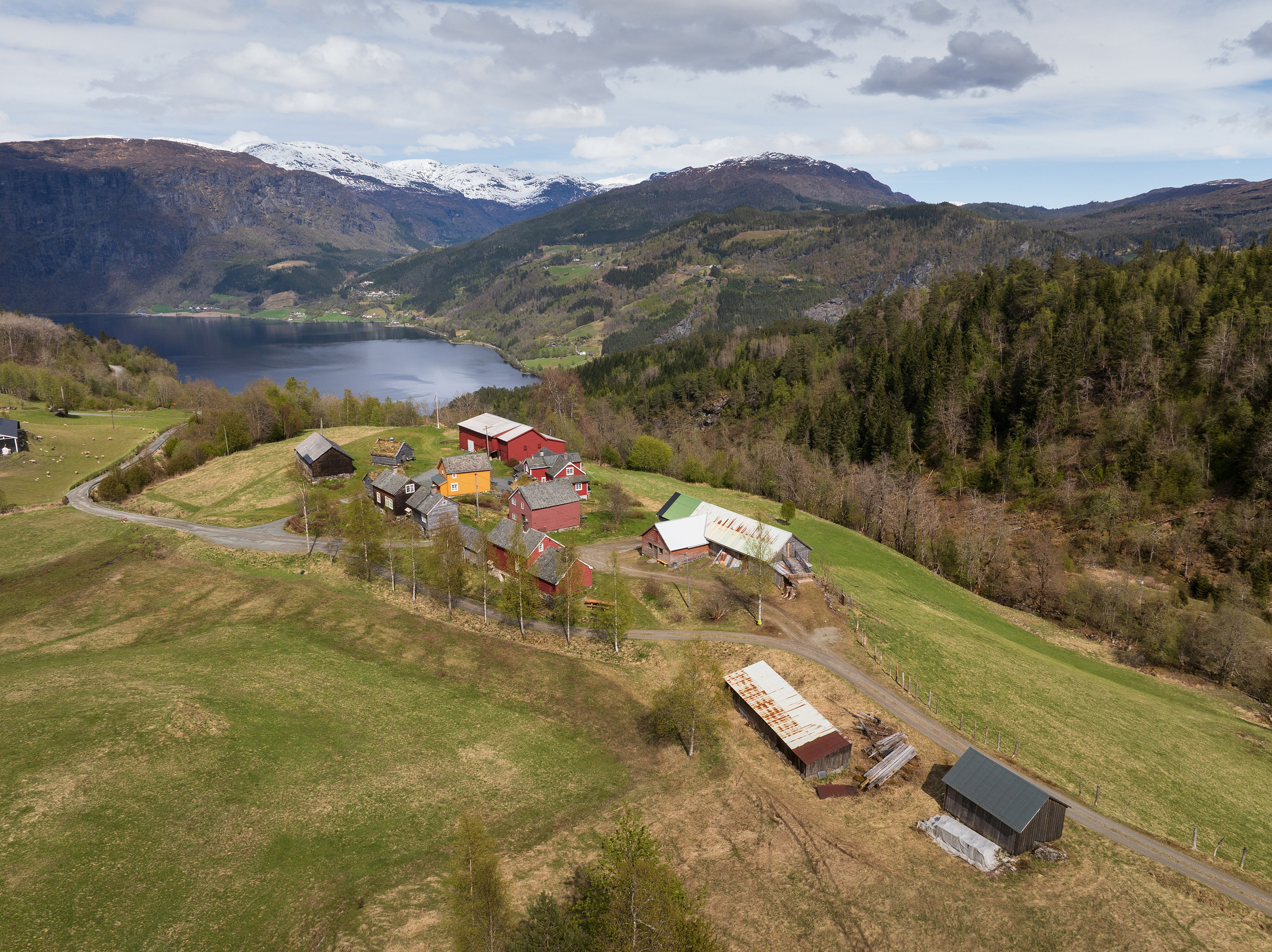 En liten klynge gårdsbygninger ligger i en gressbevokst åsside med en innsjø, skog og snødekte fjell i bakgrunnen under en delvis overskyet himmel.