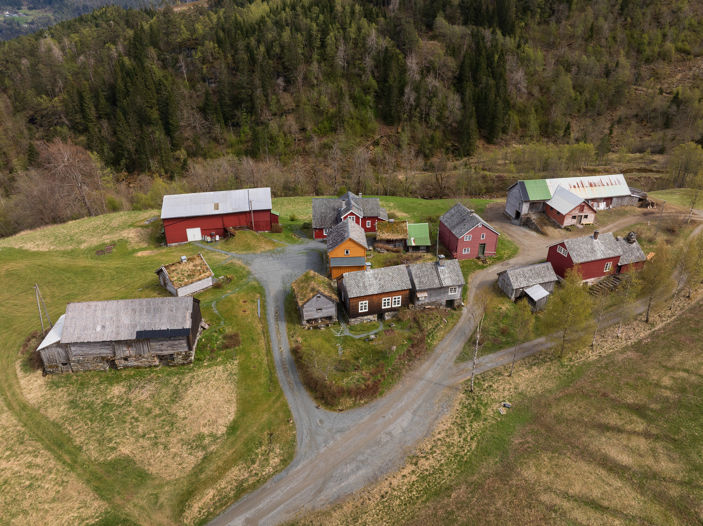 Flyfoto av en landsby med flere hus, låver og uthus omgitt av gress og skogkledde åser.