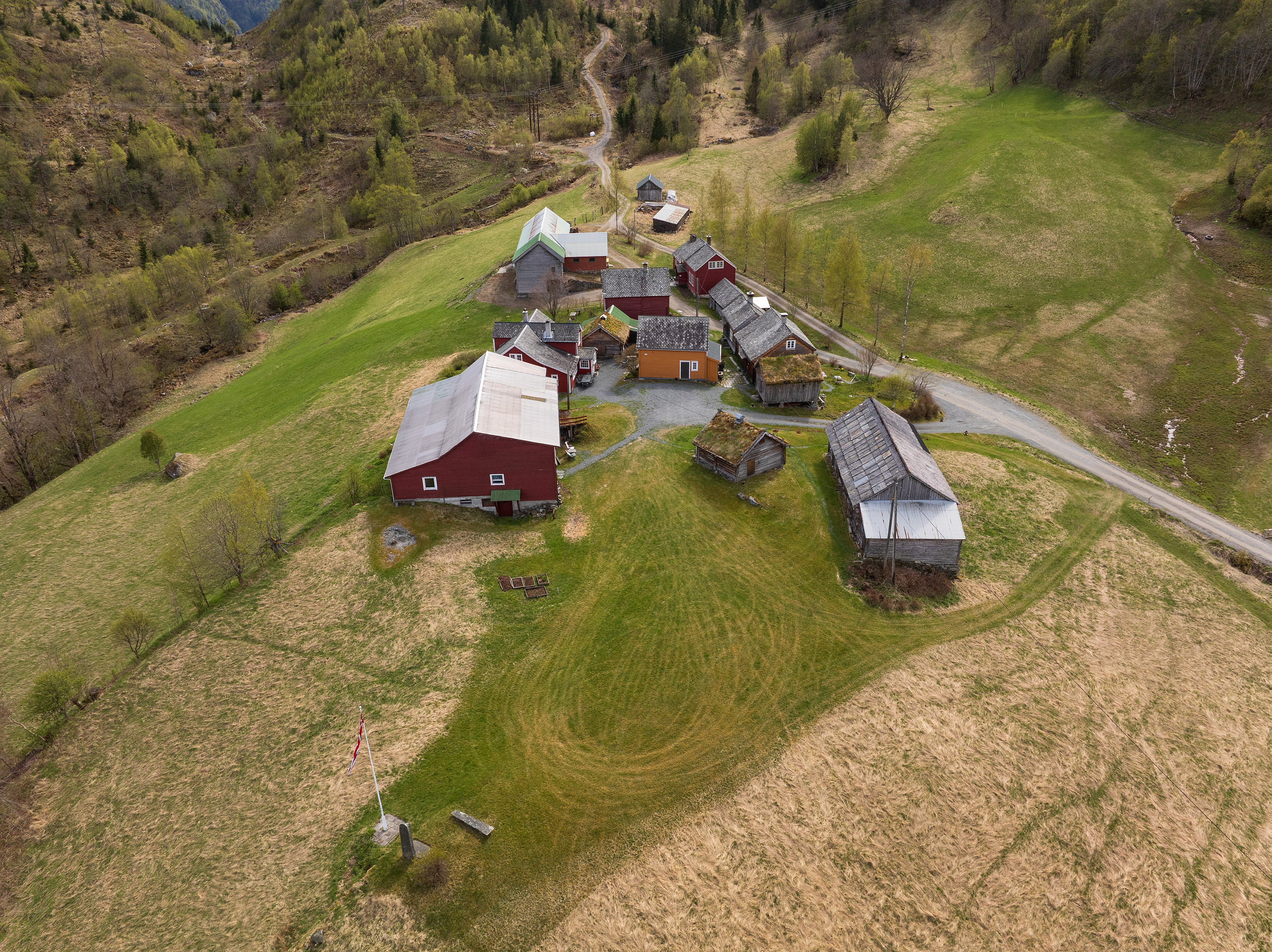 Flyfoto av en liten landsby med flere trebygninger, omgitt av grønne åkrer og trær, gjennomskåret av en smal vei.