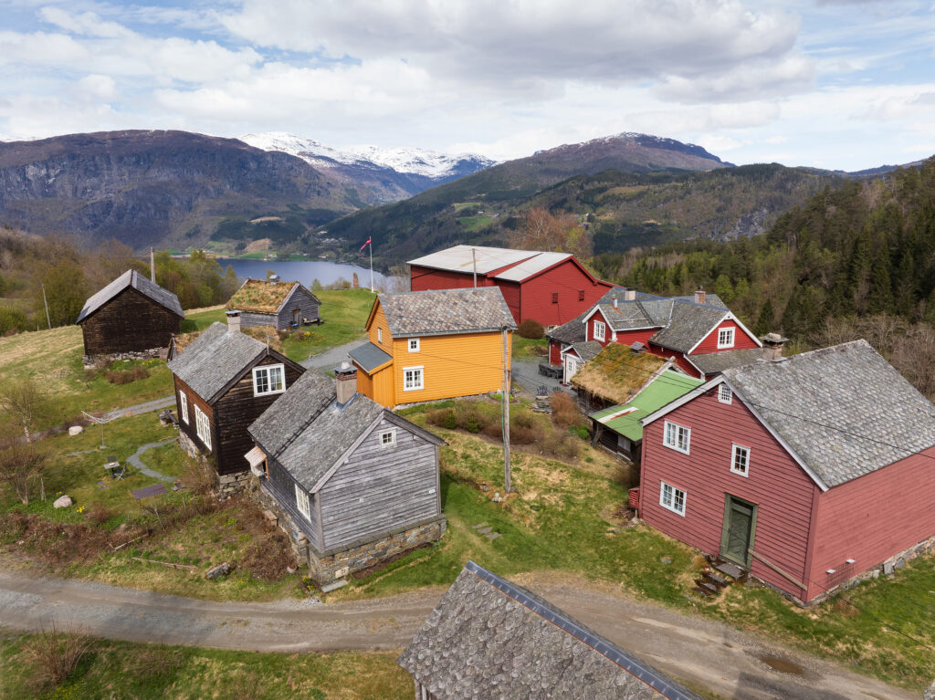 En klynge fargerike trehus ligger i en åsside omgitt av fjell og en innsjø under en delvis overskyet himmel.