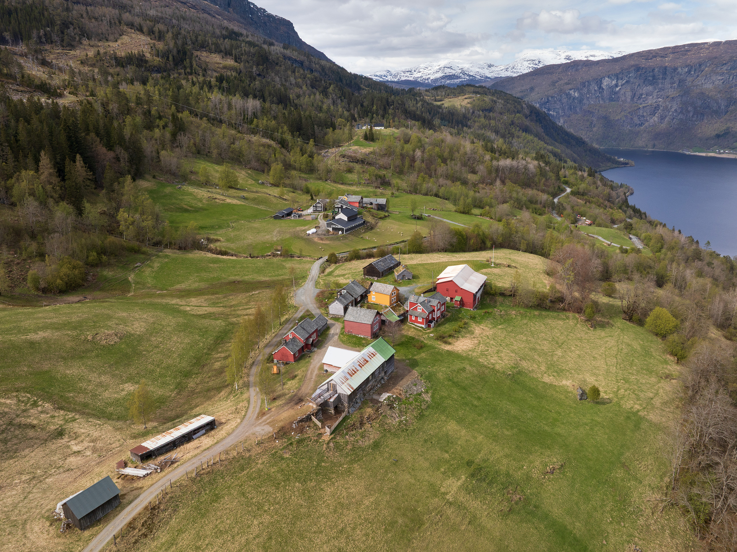 Flyfoto av en landsby med spredte hus, låver og jordbruksland, omgitt av åser, skog og en innsjø i bakgrunnen.