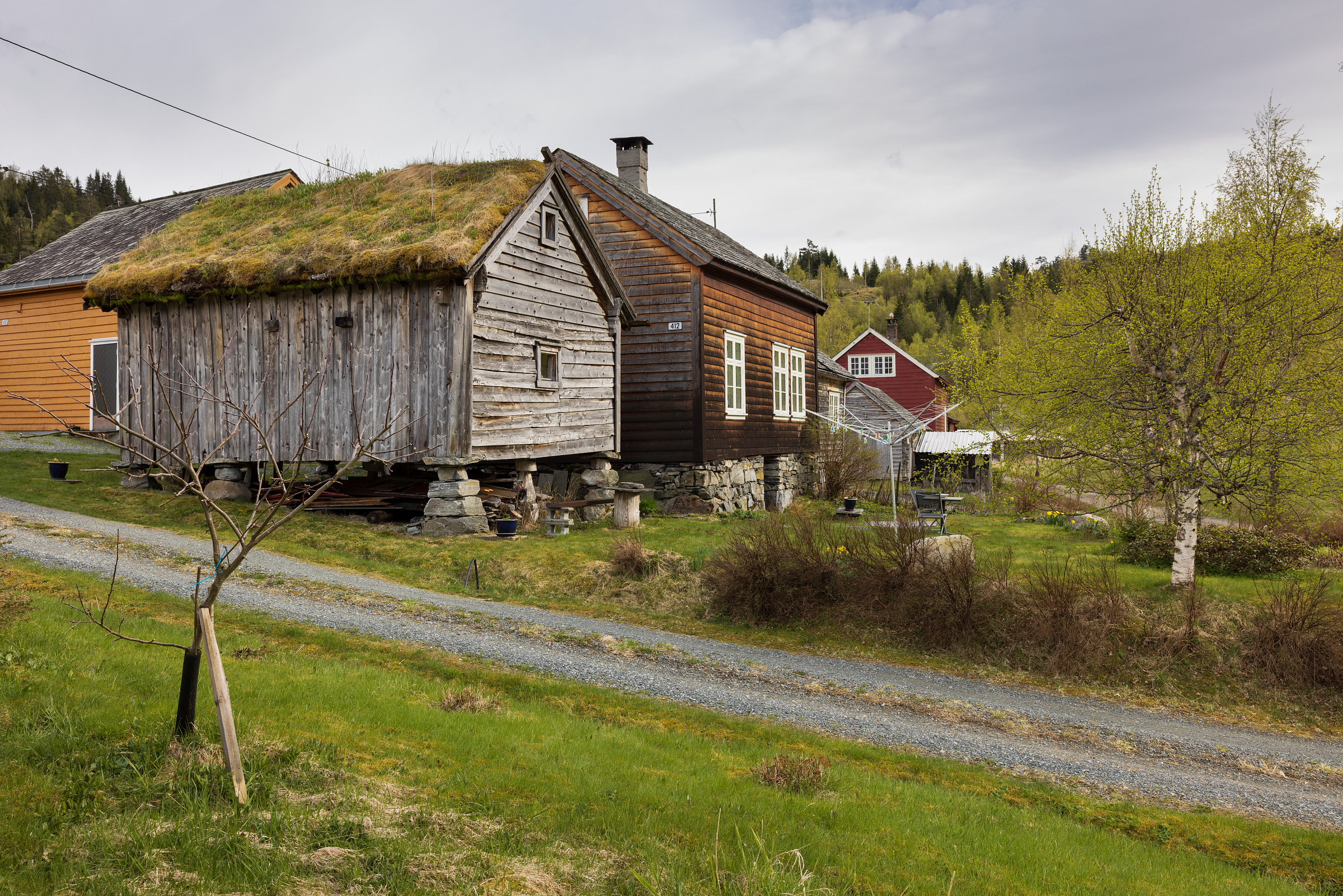 Et rustikt trehus med gressdekket tak ligger ved siden av en grusvei, omgitt av trær og flere gårdsbygninger i et landlig landskap.