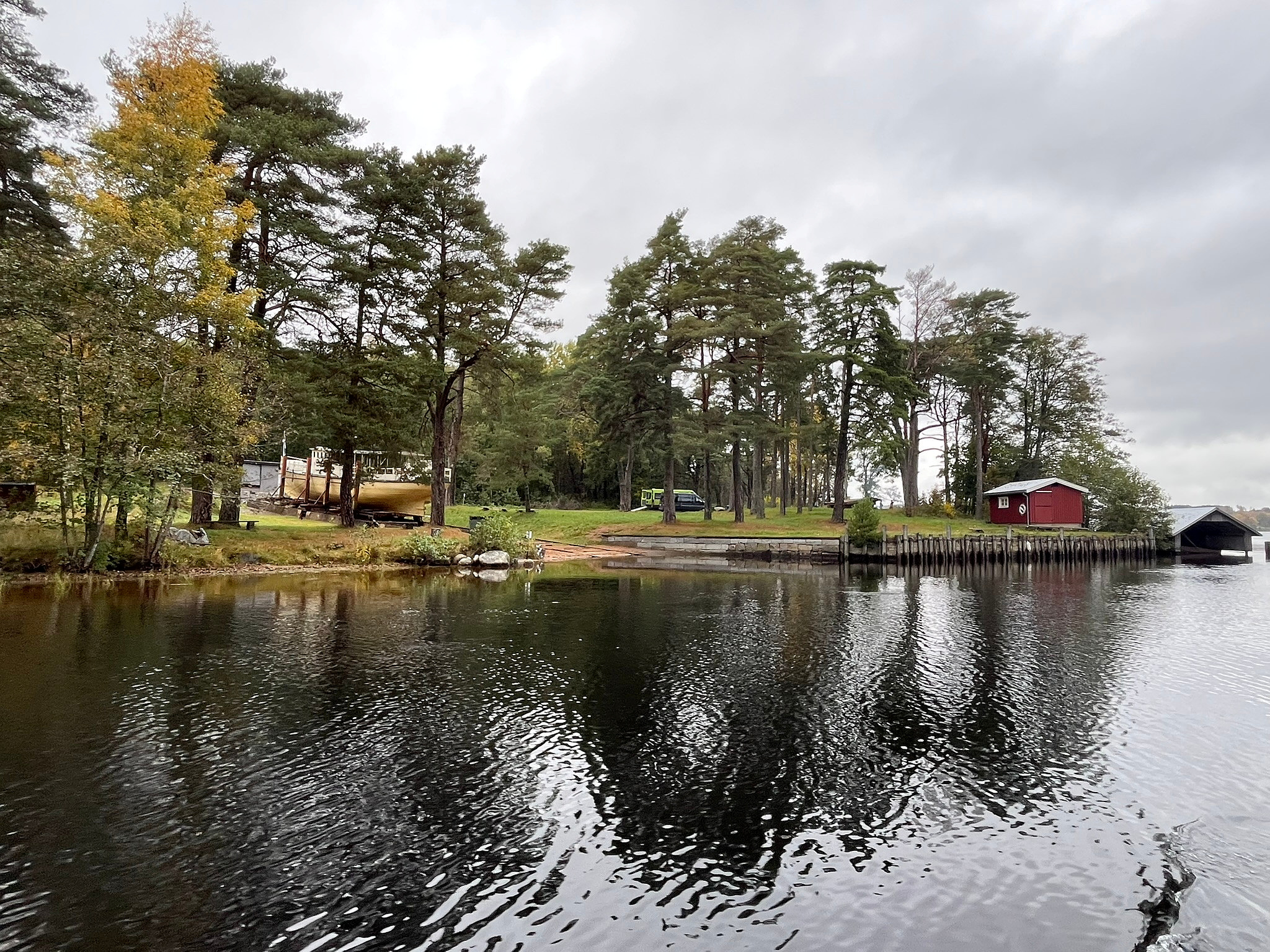 En rolig scene ved innsjøen med trær, en liten rød hytte, en båt på land og refleksjoner i vannet under en overskyet himmel.