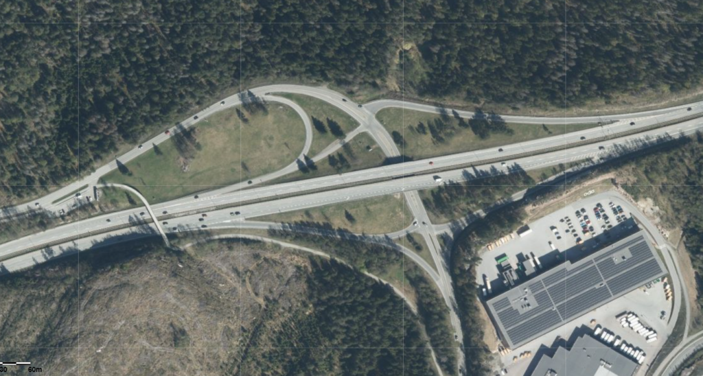 Flyfoto av et motorveikryss omgitt av skog, med en industribygning og parkeringsplass i nedre høyre hjørne.