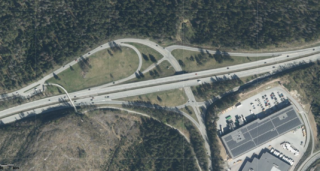 Flyfoto av et motorveikryss omgitt av skog, med en industribygning og parkeringsplass i nedre høyre hjørne.