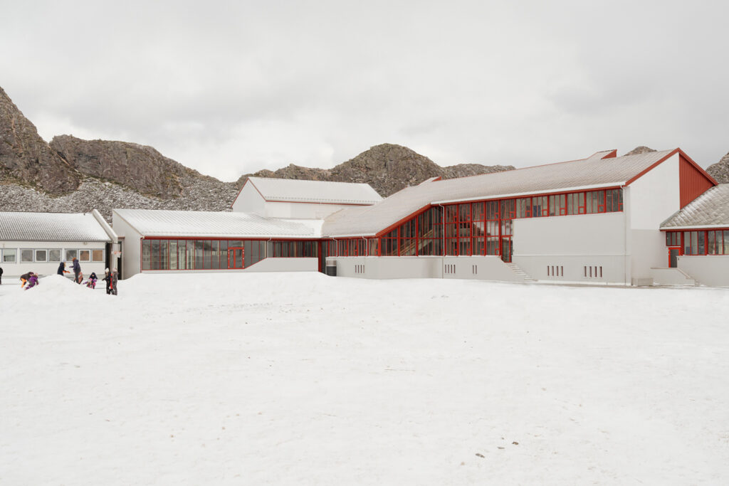 En moderne, hvit skolebygning med røde vindusrammer står i et snødekt landskap, omgitt av steinete åser. Noen få barn leker ute i nærheten av bygningen.