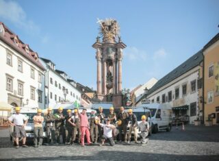 En gruppe mennesker i arbeidsuniformer og hjelmer poserer foran et barokksøylemonument på et torg i en europeisk by med omkringliggende bygninger og parkerte varebiler.