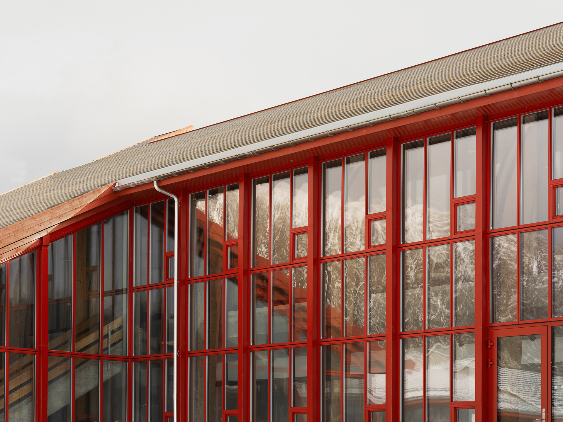 En moderne bygning med store glassvinduer innrammet i rødt, som reflekterer snødekte fjell og skyet himmel i bakgrunnen.