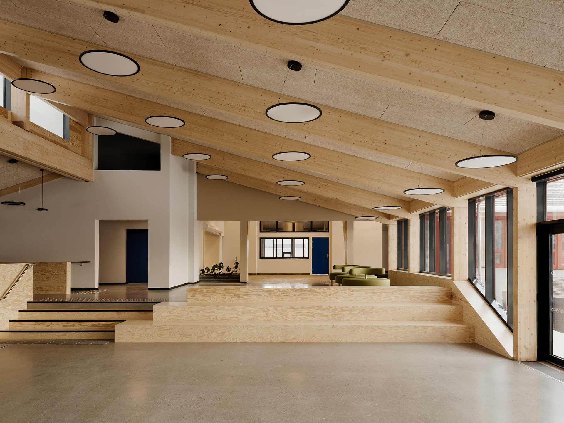 Moderne interiør med skrå tretak, runde taklamper, store vinduer og sittegrupper i lyst tre som skaper et åpent, minimalistisk rom.