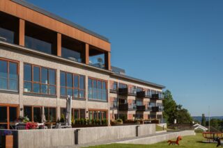 Moderne leilighetskompleks med store vinduer og balkonger, beliggende ved siden av en gressplen med uteservering og lekeapparater, under en klar blå himmel.