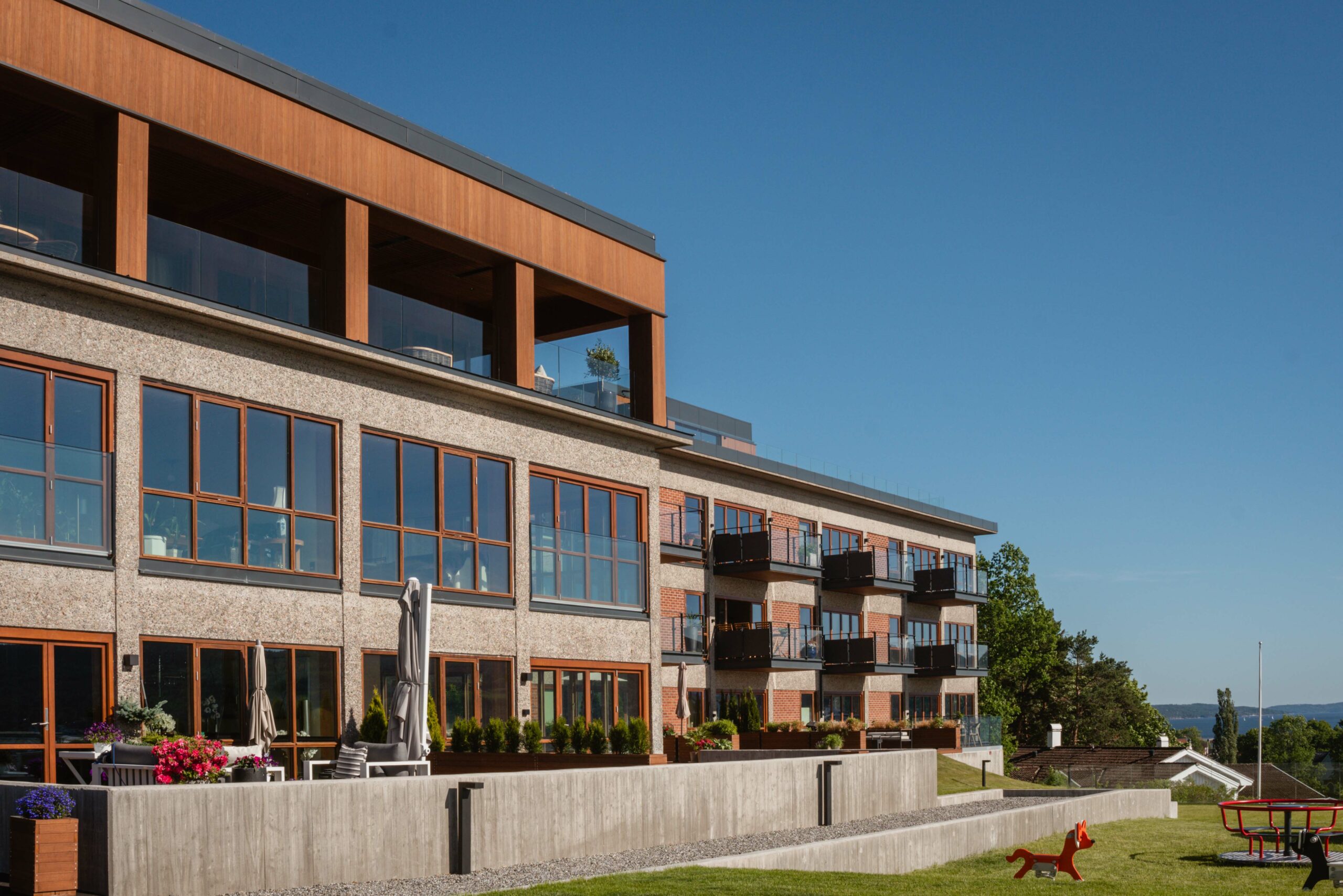 Statens arkitekturpris til Drøbak-prosjekt 1 Moderne leilighetskompleks med store vinduer og balkonger, beliggende ved siden av en gressplen med uteservering og lekeapparater, under en klar blå himmel.