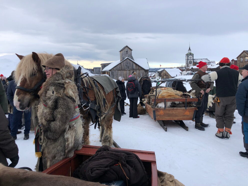 Folk samles i et snødekt landsbylandskap med hester, sleder og rustikke trehus; noen av dem har på seg vinterklær og pelsluer.