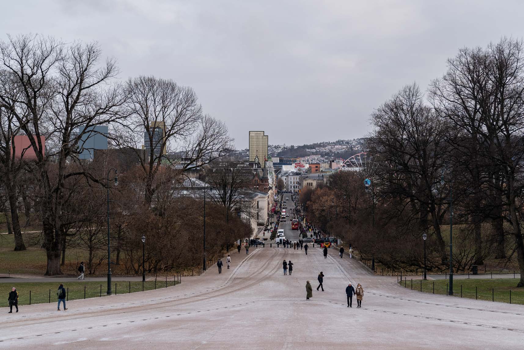 En bred, asfaltert gangvei med nakne trær på begge sider, hvor folk går, og et bylandskap synes i bakgrunnen under en overskyet himmel.