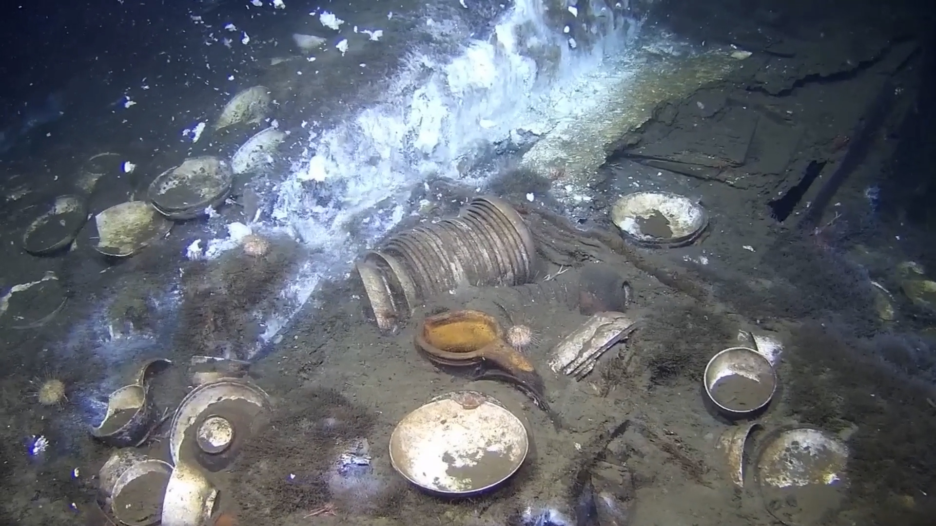 Various metal plates, bowls, and containers scattered on the seafloor, partially covered by sediment, with a cloud of particles suspended in the water above them.
