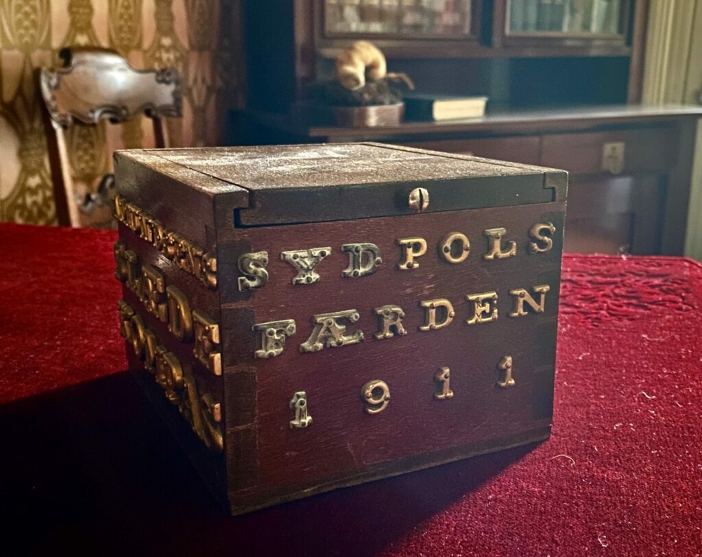 En trekasse merket SYDPOLS FÆRDEN 1911 står på en rød duk i et vintagerom med bokhyller og utsmykkede tapeter i bakgrunnen.