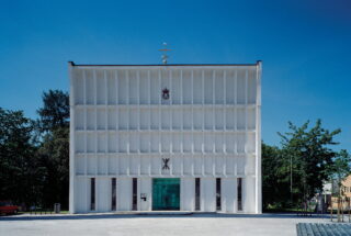 Steinkjer kirke, en moderne hvit kirke, har en symmetrisk fasade, vertikale vindusspalter og en grønn inngangsdør, som skiller seg ut mot den klare blå himmelen.