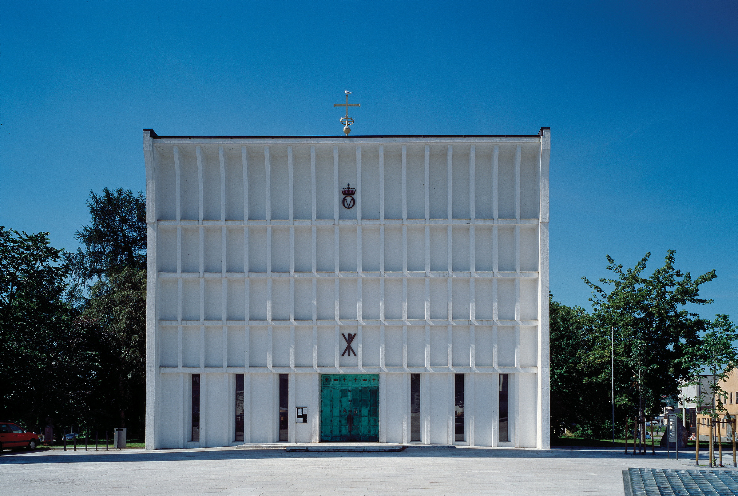 Steinkjer kirke, en moderne hvit kirke, har en symmetrisk fasade, vertikale vindusspalter og en grønn inngangsdør, som skiller seg ut mot den klare blå himmelen.
