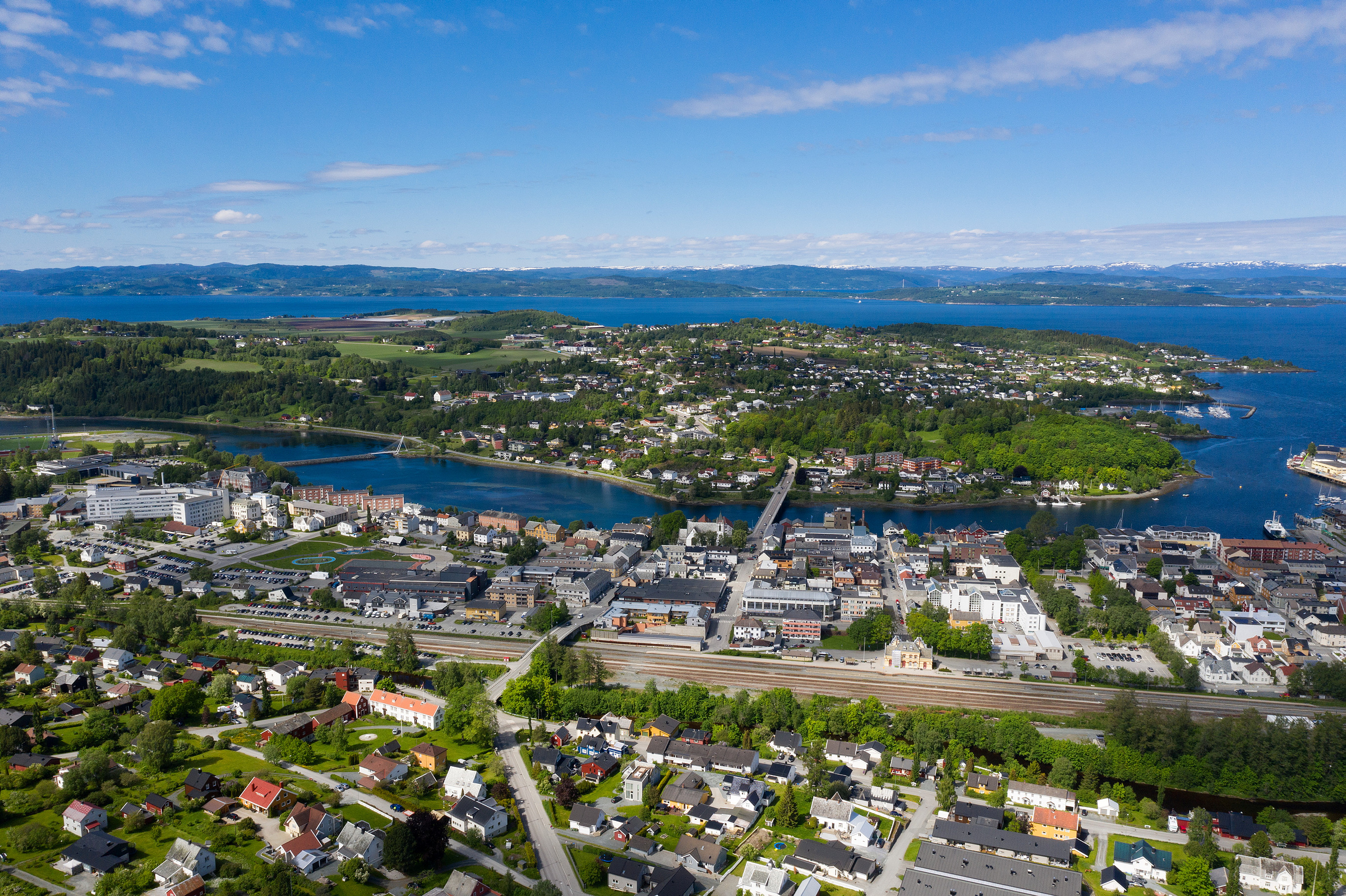 Luftfoto av en kystby med bolighus, næringsbygg, grønne områder og et vann under en blå himmel med skyer.