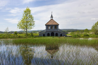 En liten, toetasjes grå paviljong med rødt tak står ved en dam, omgitt av grønt gress og trær, og speilbildet av den vises i det rolige vannet som et fredfullt bilde av et Kirke-landskap.
