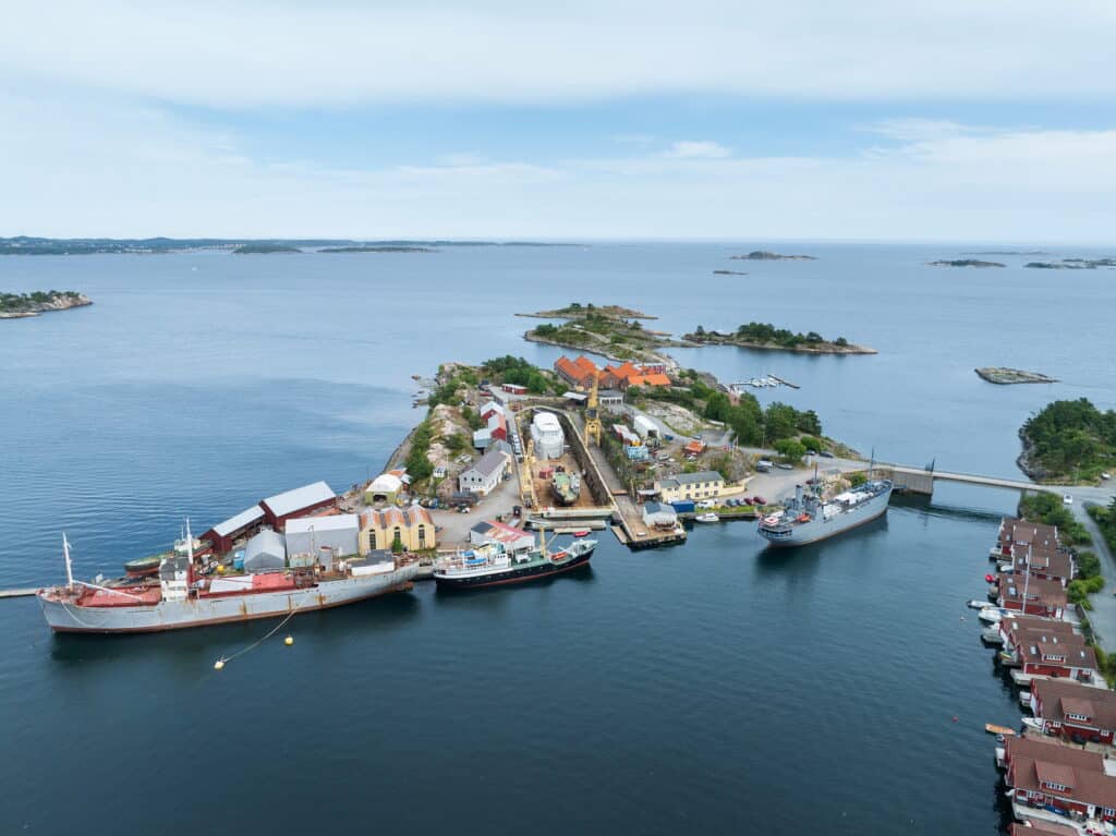 Luftfoto av Bredalsholmen Dokk og Fartøyvernsenter, en liten kystøy med kaier, flere skip, industribygg og husgrupper, omgitt av rolig, blått vann og mindre øyer i nærheten.