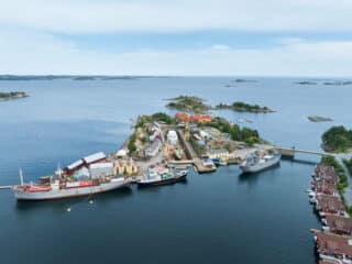 Luftfoto av Bredalsholmen Dokk og Fartøyvernsenter, en liten kystøy med kaier, flere skip, industribygg og husgrupper, omgitt av rolig, blått vann og mindre øyer i nærheten.