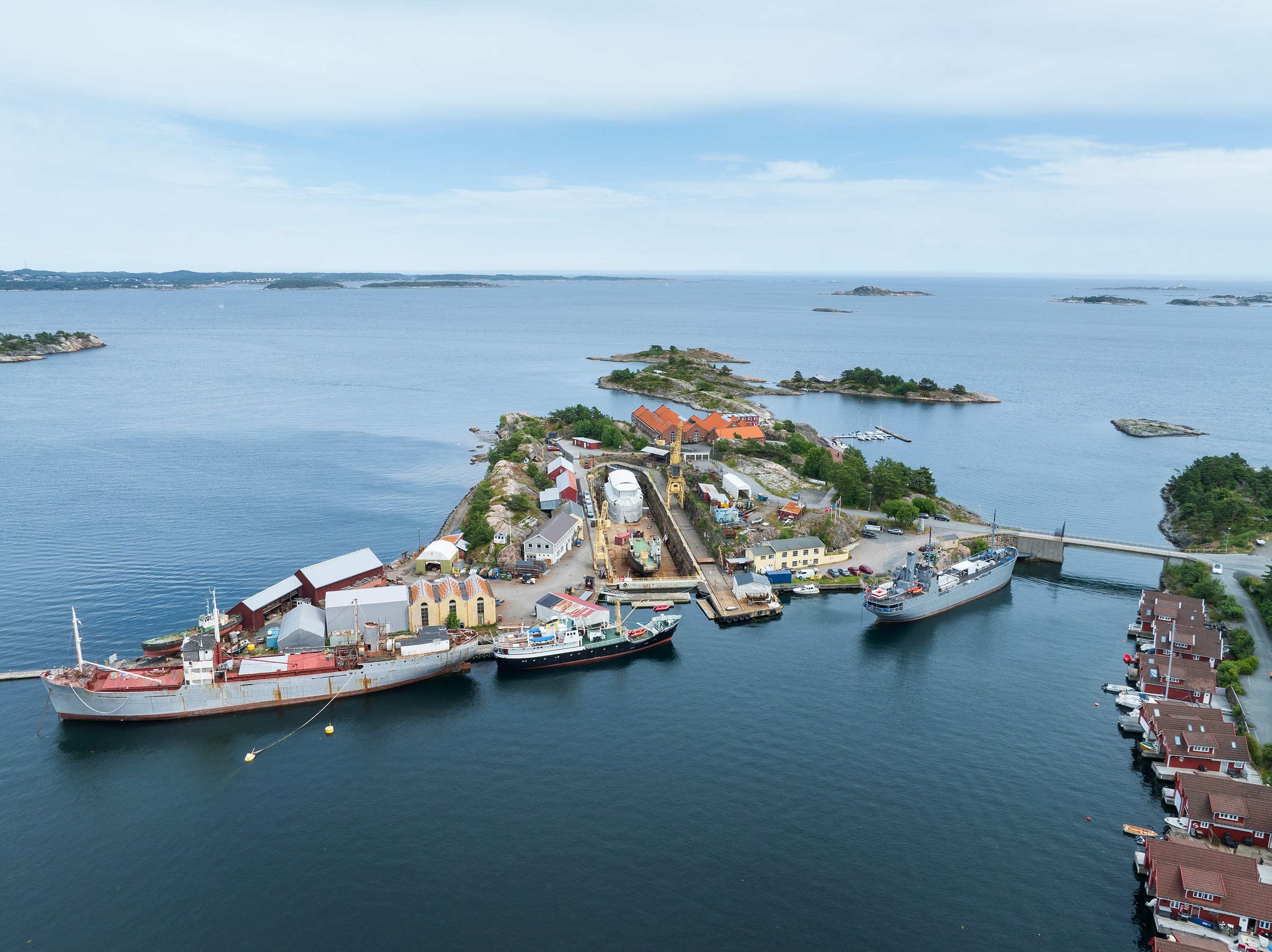 Luftfoto av Bredalsholmen Dokk og Fartøyvernsenter, en liten kystøy med kaier, flere skip, industribygg og husgrupper, omgitt av rolig, blått vann og mindre øyer i nærheten.