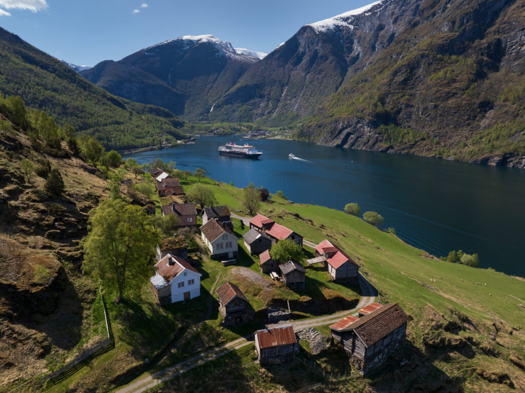 Luftfoto av en liten landsby ved en fjord, med et cruiseskip og en båt på vannet, og grønne åser og snødekte fjell i bakgrunnen – et malerisk landskap som fremheves av en historisk middelalderkirke.