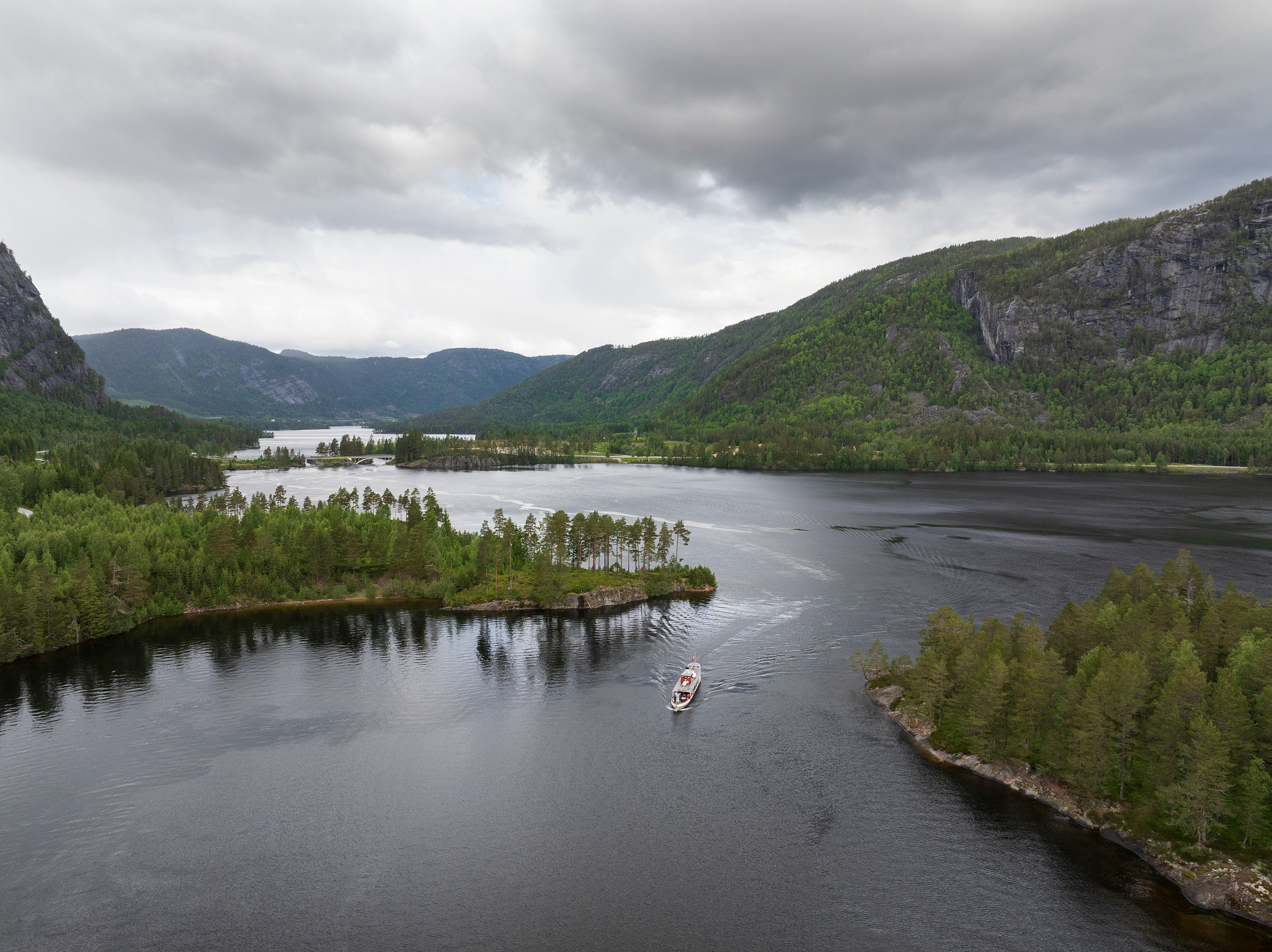 D/S Bjoren seiler på Byglandsfjorden omgitt av grønne skoger og åser under en overskyet himmel.
