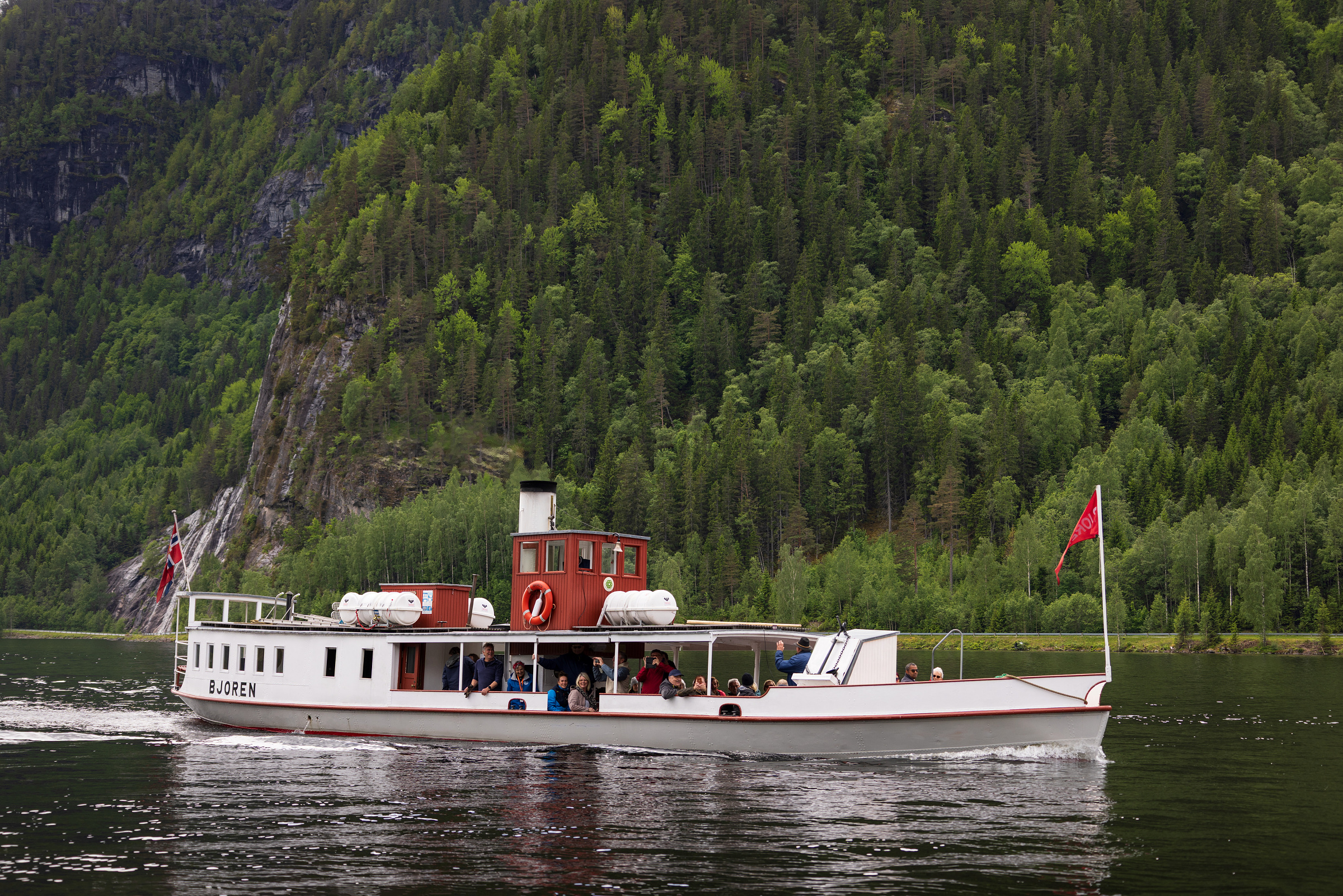 No blir D/S «Bjoren» freda 6 D/S Bjoren, en hvit passasjerbåt, med mennesker om bord beveger seg på en Byglandsfjorden, omgitt av tett, grønn skog og bratte åser.